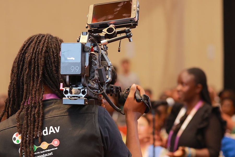 A videographer with dreadlocks filming a speaker at a corporate event using a professional cinema camera and monitor.
