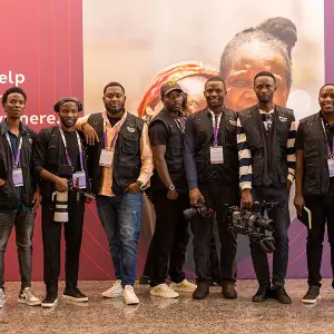 A team of professional event photographers and videographers in black vests posing with cameras in front of a purple banner.