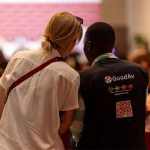 A GoodAv technician in a branded vest assisting a woman at a corporate event or conference.