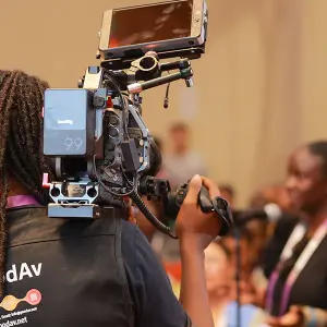 A videographer with dreadlocks filming a speaker at a corporate event using a professional cinema camera and monitor.